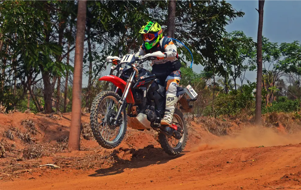 Motorcyclist in vibrant gear performing an off-road jump on a dirt track, surrounded by trees and dusty terrain during GT bike race training.
