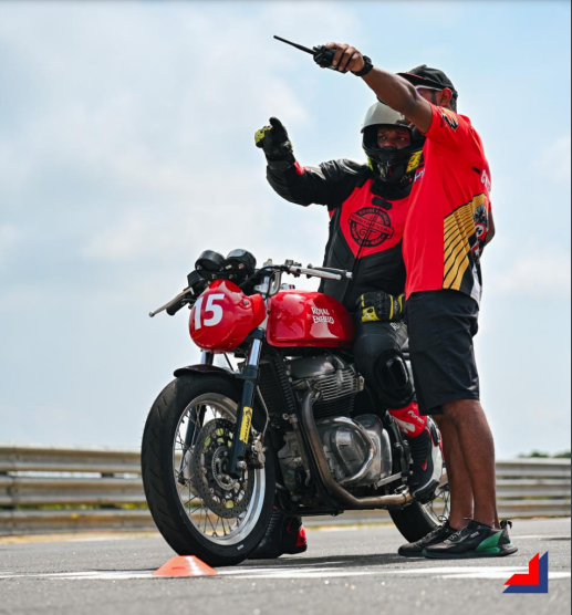 Professional motorcycle coaching session with a rider on a red Royal Enfield, guided by an expert coach during a road race training event.