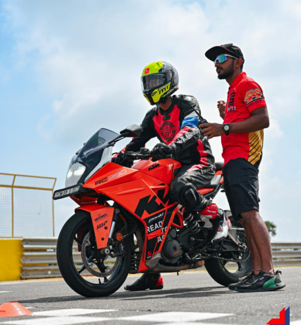 Motorcycle racer on an orange KTM RC 390 bike receiving instructions from a trainer on a race track.